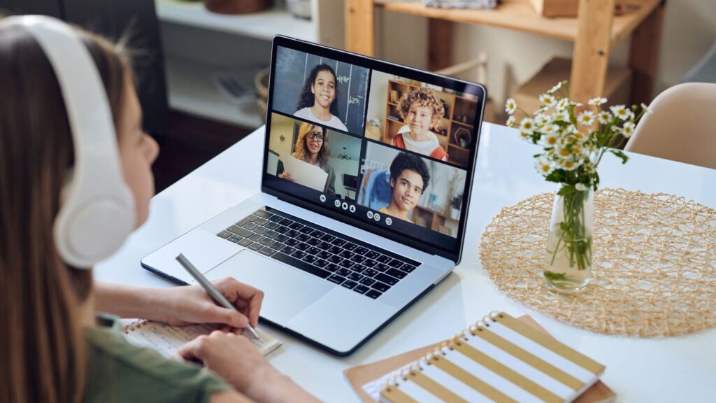 Girl with headphones participating in a small group online reading tutoring session with certified teacher and students on laptop screen